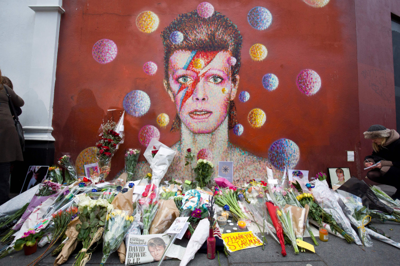 Floral tributes are left beneath a mural of British singer David Bowie, painted by Australian street artist James Cochran, aka Jimmy C, following the announcement of Bowie's death, in Brixton, south London, on January 11, 2016. British music icon David Bowie died of cancer at the age of 69, drawing an outpouring of tributes for the innovative star famed for groundbreaking hits like "Ziggy Stardust" and his theatrical shape-shifting style. AFP PHOTO / JUSTIN TALLIS RESTRICTED TO EDITORIAL USE, MANDATORY MENTION OF THE ARTIST UPON PUBLICATION, TO ILLUSTRATE THE EVENT AS SPECIFIED IN THE CAPTION / AFP / JUSTIN TALLIS (Photo credit should read JUSTIN TALLIS/AFP/Getty Images)