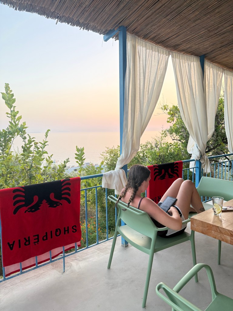 A person sitting on a balcony with a view of the sea, reading a book. The balcony is adorned with sheer curtains and towels with an emblem, surrounded by greenery and a sunset sky.