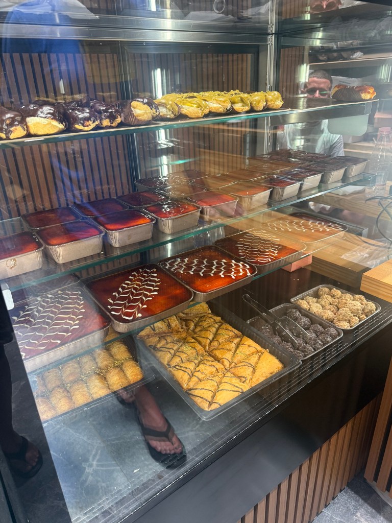 A display case filled with various desserts, including pastries topped with chocolate, yellow cream-filled cones, and trays of layered sweets, showcasing an array of colors and textures.