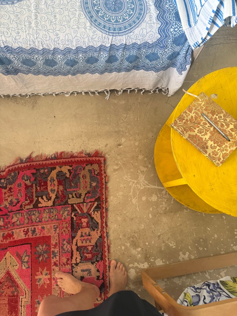 A cozy indoor setting with a colorful patterned rug, a blue and white bedspread, a yellow round table with a notebook, and a person's bare feet visible.