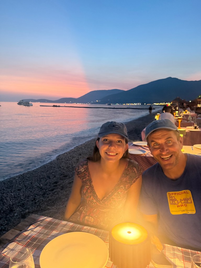 A couple smiling at a beachside restaurant during sunset, with a candle on the table and mountains in the background.