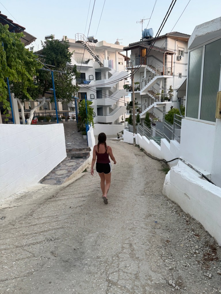 A person walking down a narrow pathway in a residential area, flanked by white walls and buildings with multiple staircases.