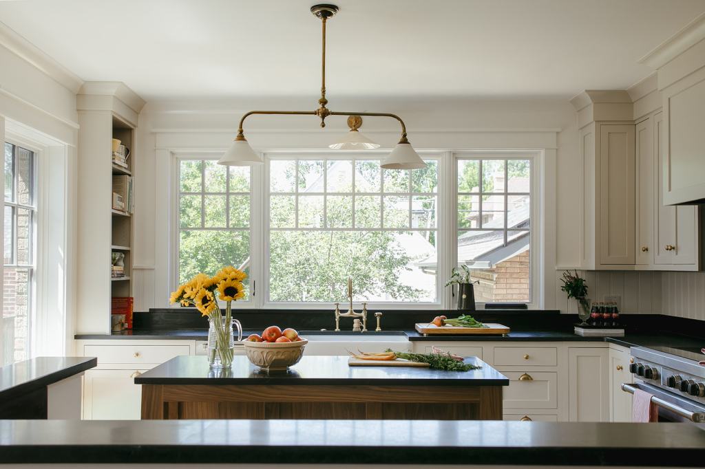 A bright and modern kitchen featuring a large window with a view of greenery, a black countertop with fresh vegetables and fruit, and a vase of sunflowers on the counter under pendant lighting.