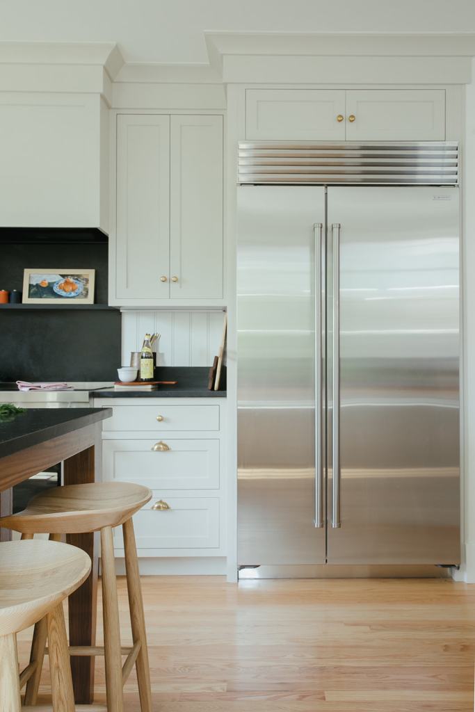 A modern kitchen featuring a stainless steel refrigerator, white cabinets with gold handles, and a dark countertop. Two wooden bar stools are visible in the foreground.