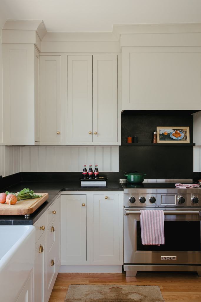 Modern kitchen with white cabinets, black countertops, and stainless steel appliances including a Wolf stove. A wooden cutting board with vegetables is on the counter, alongside a small display of Coca-Cola bottles and a green pot.