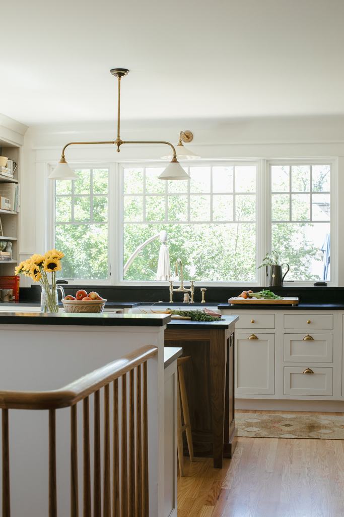 Bright and modern kitchen featuring a large window with greenery outside, a central island with a black countertop, wooden bar stools, and a vase of sunflowers on the countertop.