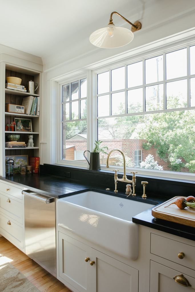 Modern kitchen featuring a farmhouse sink, stainless steel dishwasher, and a black countertop, illuminated by a decorative wall sconce and large windows providing natural light.