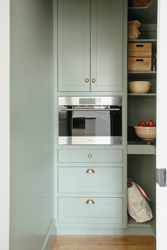 A modern kitchen pantry showcasing mint green cabinetry, featuring a built-in microwave and multiple storage drawers. Wooden shelves hold a bowl of apples and woven storage baskets, with a white tote bag visible on the lower shelf.